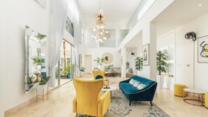 A spacious living area with a yellow armchair, a blue sofa, and a round coffee table. There are multiple potted plants and artwork on the walls.