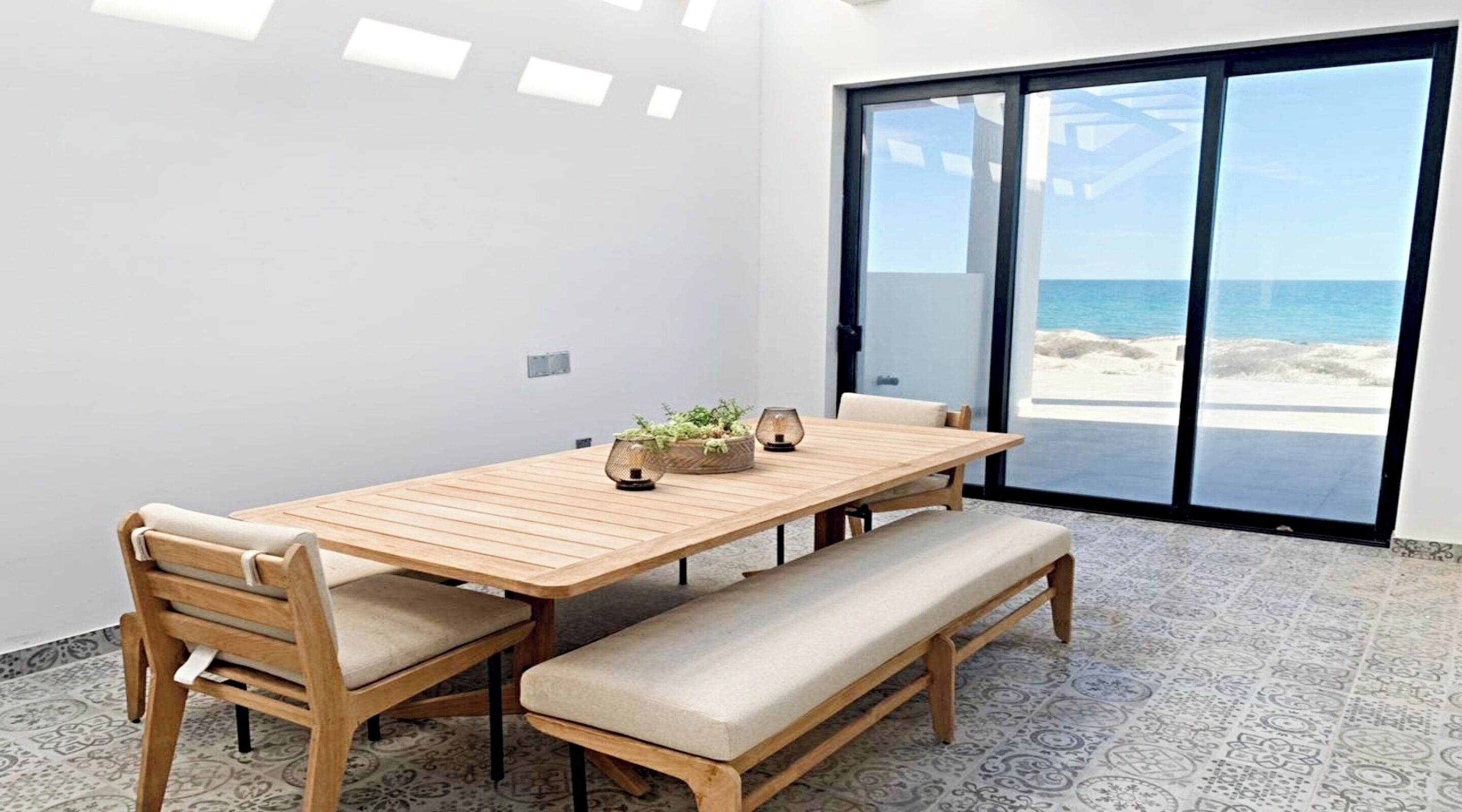 A dining area with a wooden table, cushioned chairs, and a bench. It features a large glass door leading to a beach view.