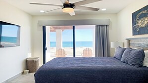 A bedroom with a large bed, a ceiling fan, a view of the beach, and a framed artwork on the wall.