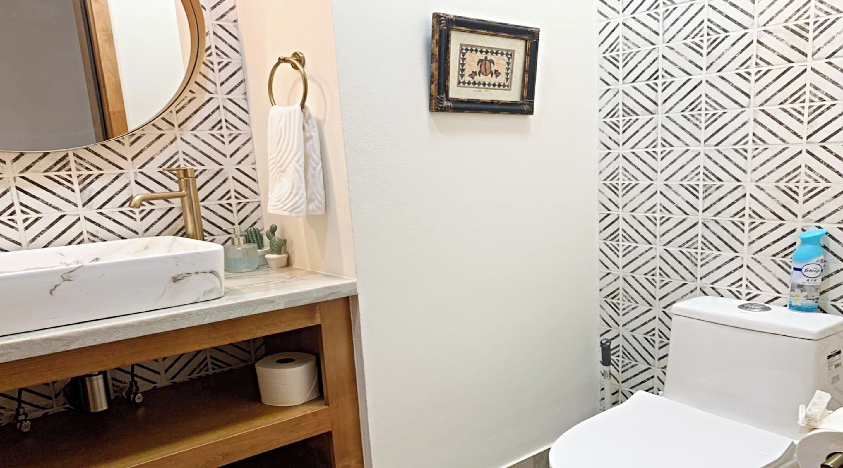 Un baño con lavamanos blanco, grifo dorado, espejo, un cuadro enmarcado y azulejos estampados.