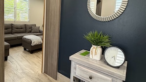 A hallway with a dark wall, a round mirror, a small wooden console table with a clock and a plant, and a view into a living room with a sofa and a window. - EcoMar Rentals #4: Near Beach Couple's Tropical Retreat (Cabo Rojo)