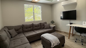 A modern living room with a sectional sofa, a flat-screen TV, and a ceiling with geometric lighting fixtures. - EcoMar Rentals #4: Near Beach Couple's Tropical Retreat (Cabo Rojo)