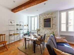 A dining area with a table set for four, surrounded by chairs, a glass shelving unit with dishes, and a rustic ceiling beam. - Appartement Tonnel - Welkeys (Bandol)