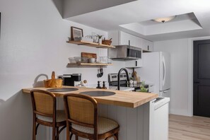 Une cuisine compacte avec un îlot en bois, deux chaises en osier, un four à micro-ondes, un réfrigérateur et un petit évier.