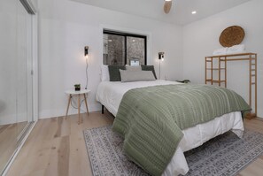Une chambre avec un lit, une petite table sur laquelle est posée une plante, une fenêtre donnant sur un mur en pierre et une échelle en bois appuyée contre le mur.
