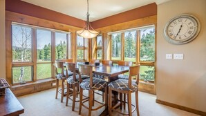 A dining area with a wooden table and chairs, large windows with a view of trees, and a wall clock. - Family Friendly w/ Suncadia Pool Access (Cle Elum)