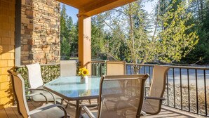 A covered patio with a glass table and metal chairs, surrounded by a stone wall and a railing overlooking a wooded area. - Pet Friendly Condo, Suncadia Pool Access (Cle Elum)