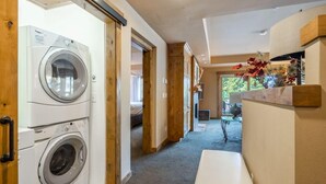 Een wasserette met twee wasmachines, een houten bankje en uitzicht op een slaapkamer met een bed en een raam.