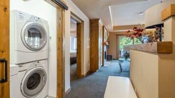 A laundry room with two washing machines, a wooden bench, and a view of a bedroom with a bed and a window.