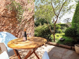 A wooden table with a bottle and glasses, set against a brick wall and surrounded by greenery.