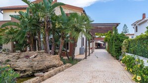 A paved pathway leading to a building with a stone facade, surrounded by tropical plants and a small rock feature. - Syracuse sea (Arenella)