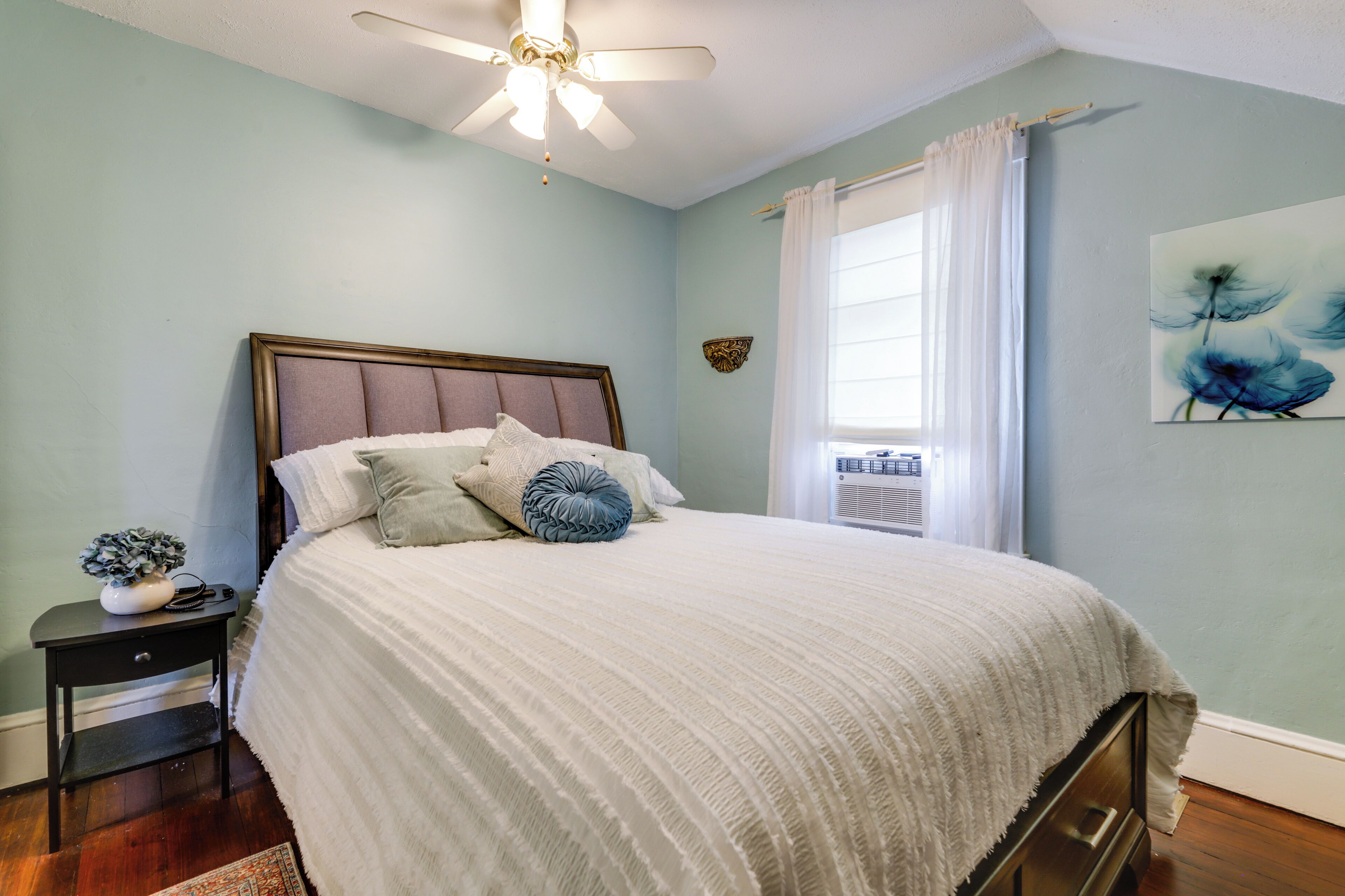 A bedroom with a bed, a nightstand, a ceiling fan, a window with curtains, and a wall-mounted air conditioning unit.