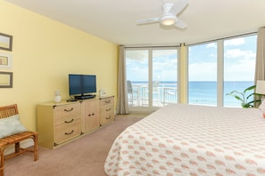 Habitación de hotel con cama, televisor, escritorio con cajones, silla y balcón con vista al océano.