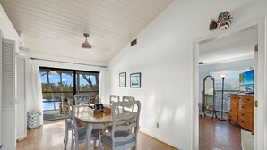 Un comedor con mesa y sillas de madera, vistas al exterior y un cuadro en la pared.