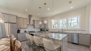 A modern kitchen with a central island, bar stools, stainless steel appliances, and large windows. - 30A Brand NEW 4 Bedroom-8 Bikes-LARGE POOL-Turf yard Close to Beach-Golf Cart! (Santa Rosa Beach)