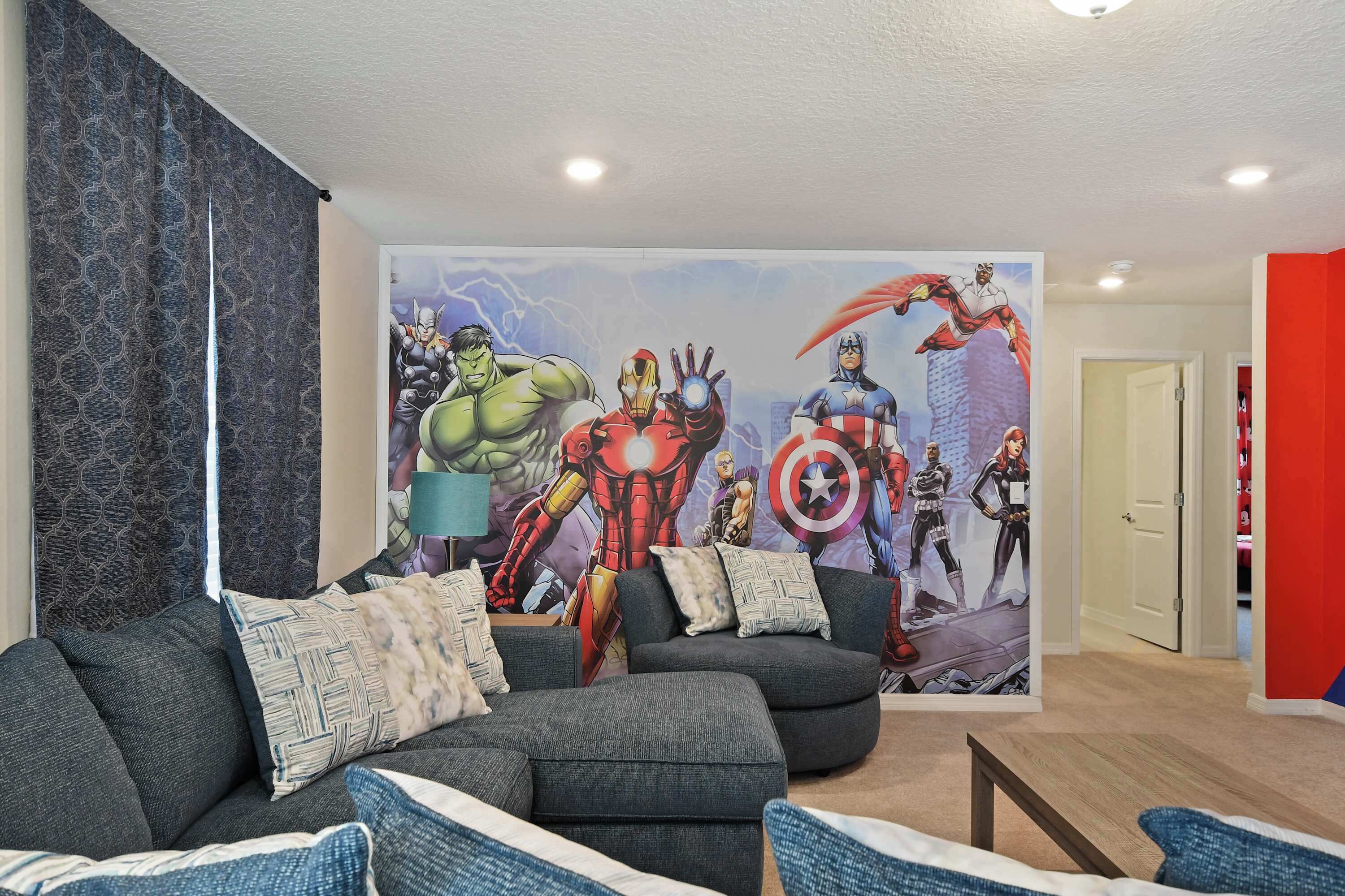 A living room with a large superhero mural, a grey sofa, and a red accent wall.