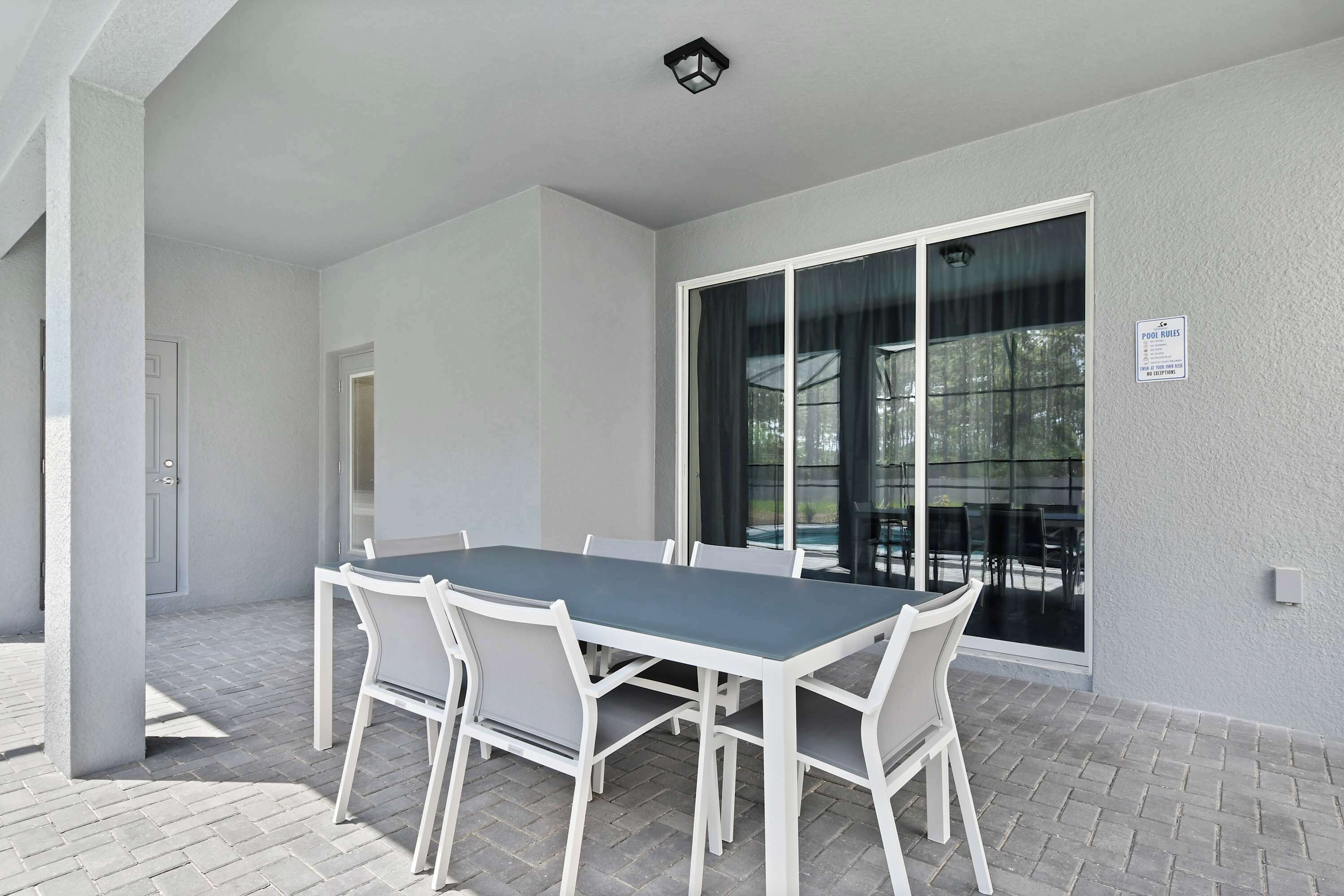 A covered patio with a table and chairs, a sliding glass door, and a sign indicating pool hours.