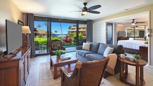 A hotel room with a balcony, a sofa, a dining table, a chair, a TV, and a ceiling fan.