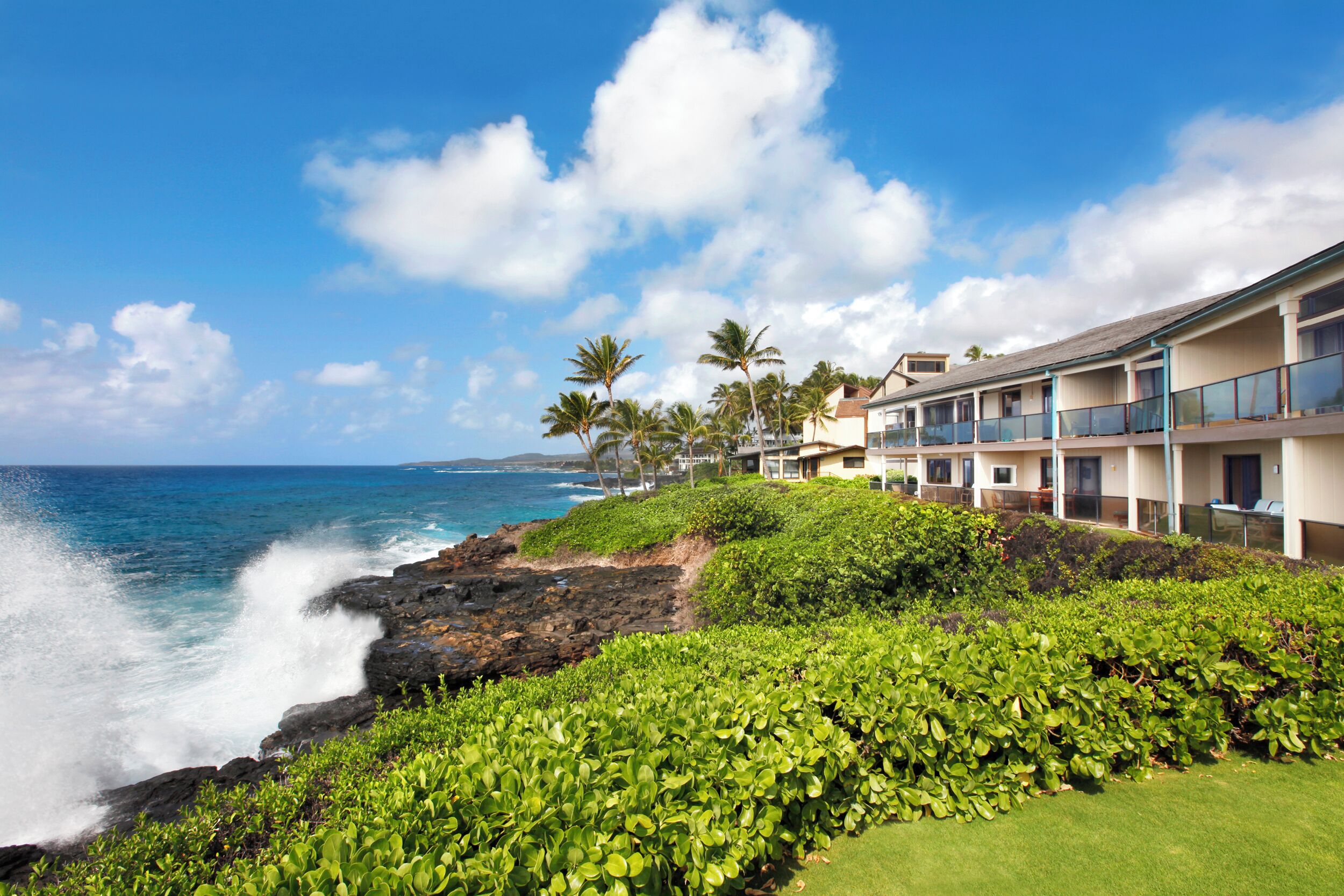 A coastal hotel with a view of the ocean, featuring multiple balconies, lush greenery, and a clear sky.