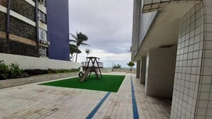 A paved walkway with a green mat and a swing set, bordered by buildings with tiled walls and a palm tree in the background. - Apto 701 em Boa Viagem  (Recife)