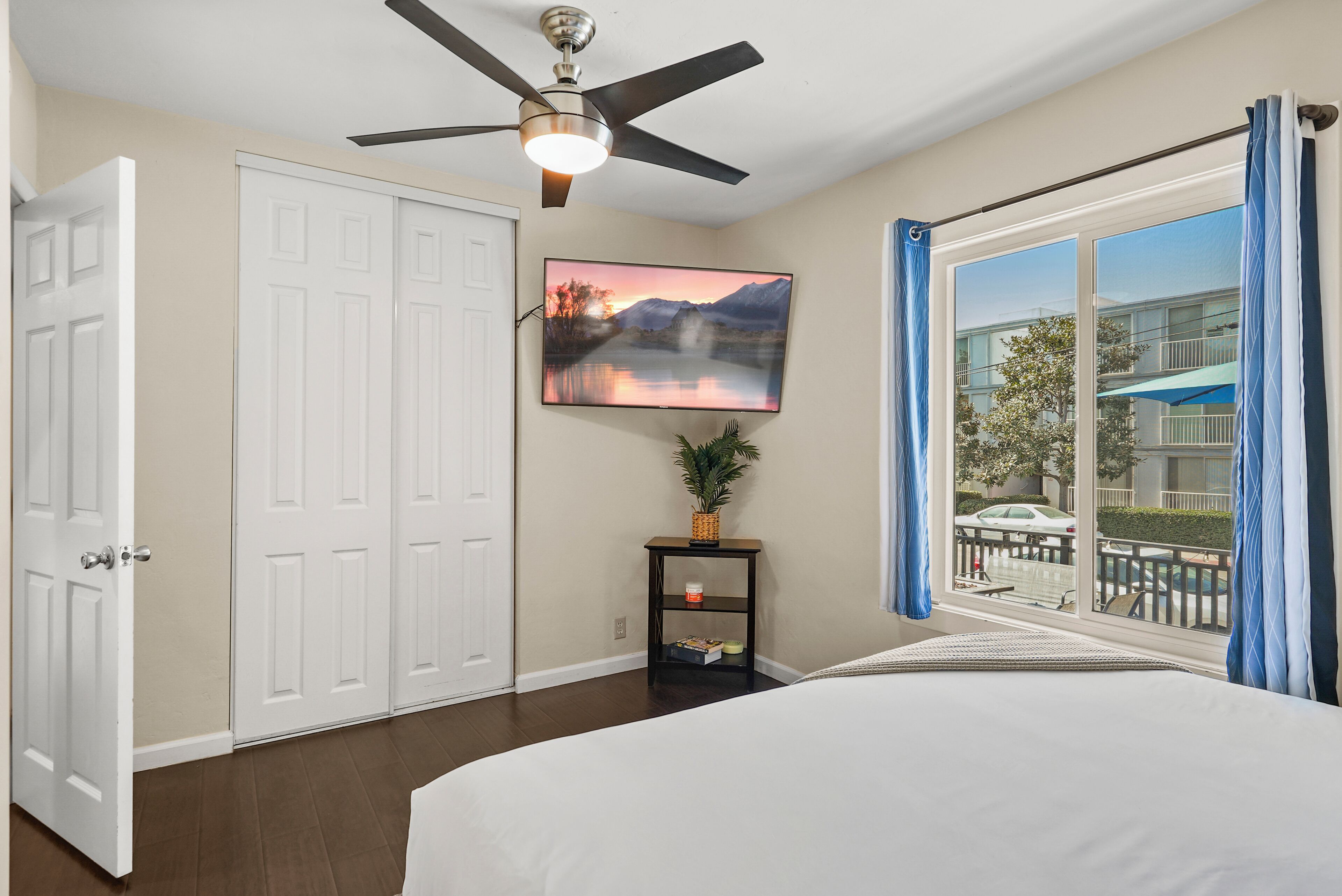 A bedroom with a bed, a ceiling fan, a TV mounted on the wall, a small table with a plant, and a window with curtains.