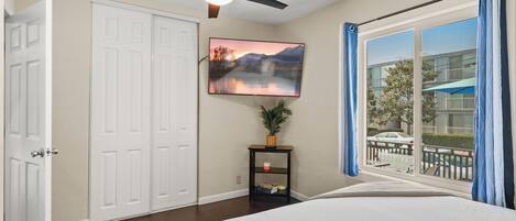 A bedroom with a bed, a ceiling fan, a TV mounted on the wall, a small table with a plant, and a window with curtains.