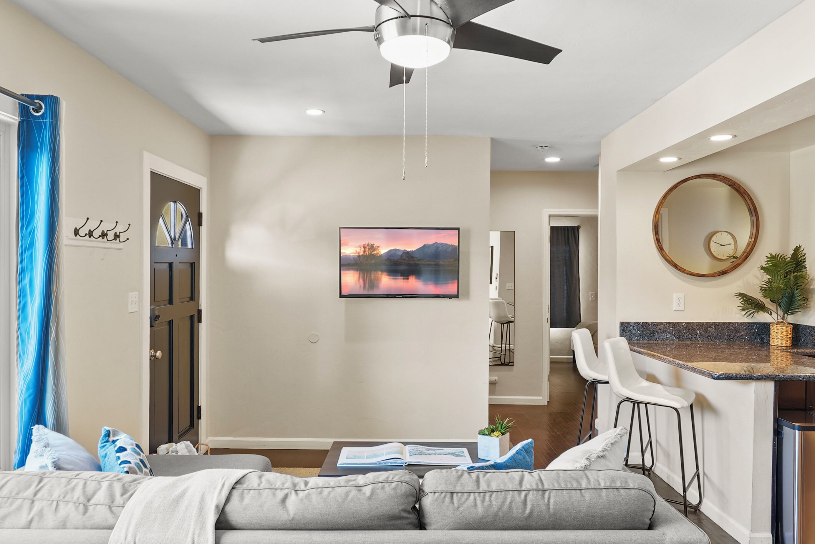 A modern living room with a gray sofa, a coffee table with a book, a TV mounted on the wall, and a ceiling fan.
