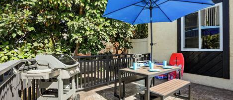 A patio with a barbecue grill, a table with chairs, and a blue umbrella.