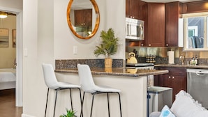 A modern kitchen with dark wood cabinets, a granite countertop, and stainless steel appliances. There are two white bar stools and a small potted plant. - Riviera Bay View Dream - Private Patio with Bay View, Parking & W/D! (San Diego)