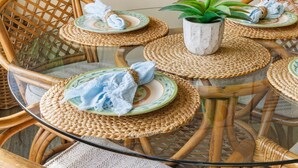 A round glass table surrounded by wicker chairs, each with a woven placemat and a plate with a blue cloth. A potted plant is placed on the table. - La Jolla Dream! Amazing Location - Large Patio, Fire Pit and Parking! (La Jolla)