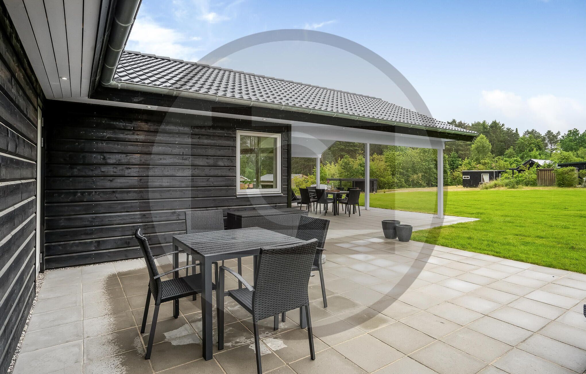 Une terrasse couverte avec une table et des chaises, donnant sur une pelouse verdoyante, et un bâtiment à l’extérieur en bois foncé.