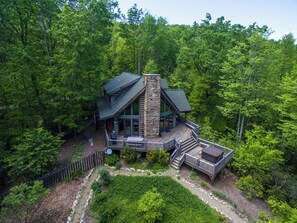 A cabin with a stone chimney, wooden deck, and surrounded by dense forest.
