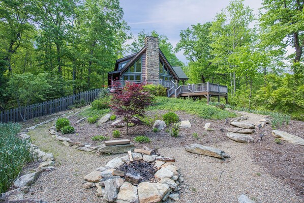 A lodge with a stone chimney, wooden deck, and a fire pit surrounded by rocks and greenery.