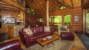 Ein Blockhaushaus-Interieur mit Holzbalkendecke, Kamin aus Stein, Ledersofa und Essbereich mit Holztischen und -stühlen.