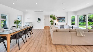 Een moderne woonkamer met een grote beige bank, een eettafel met stoelen, en een keuken die zichtbaar is door een open deur.