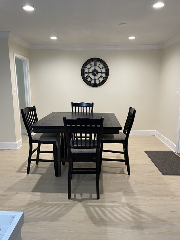 A dining area with a rectangular table, four chairs, a wall clock, recessed ceiling lights, and a door.