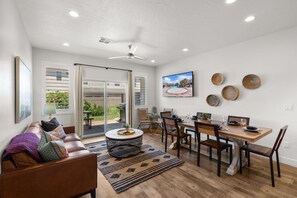 A spacious living room with a sofa, dining table, and chairs, a ceiling fan, and a large window with a view of a pool and greenery.