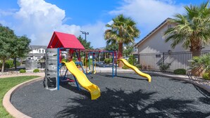 A playground with two slides and a climbing structure, surrounded by a landscaped area with palm trees and a building in the background. - Freedom Village Retreat, Neighboring units, Near St George (Washington)