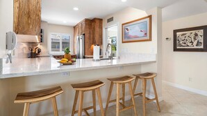 A modern kitchen with wooden cabinets, a marble countertop, and a fruit bowl. There are three wooden stools and a refrigerator.