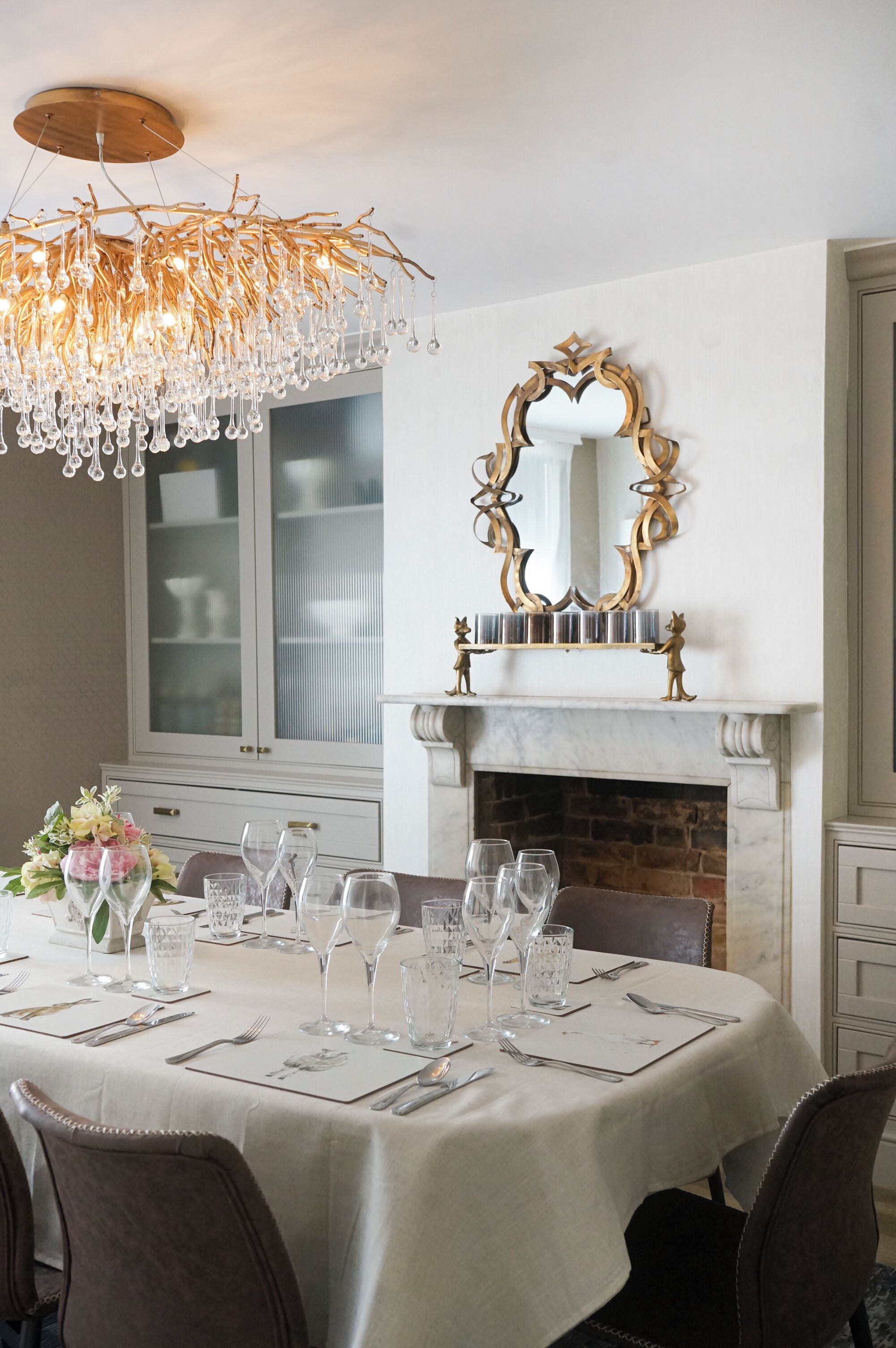 A dining room with a white table set for six, a chandelier, a fireplace, and a decorative mirror.