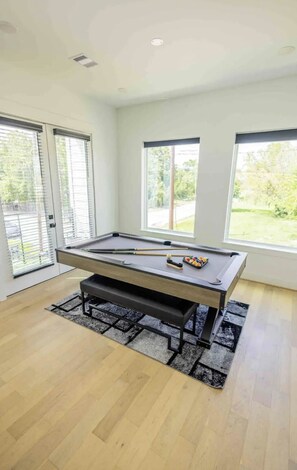 Une table de billard dans une pièce dotée de grandes fenêtres, d’un banc et offrant une vue sur la verdure à l’extérieur.