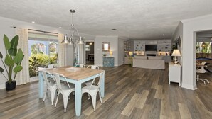 Uma sala de estar ampla com uma mesa de jantar de madeira e cadeiras brancas, uma cozinha ao fundo e uma janela grande com cortinas.