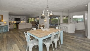 Uma cozinha espaçosa com mesa de jantar de madeira e cadeiras brancas, uma ilha grande com banquetas e vista para o exterior através de grandes janelas.