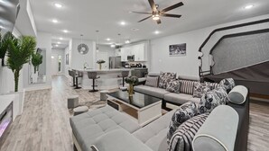 A modern living room with a sofa, coffee table, and a dining area with bar stools. - Modern 2024 Mansion with Indoor Basketball, Huge TVs & Near Downtown (Houston)