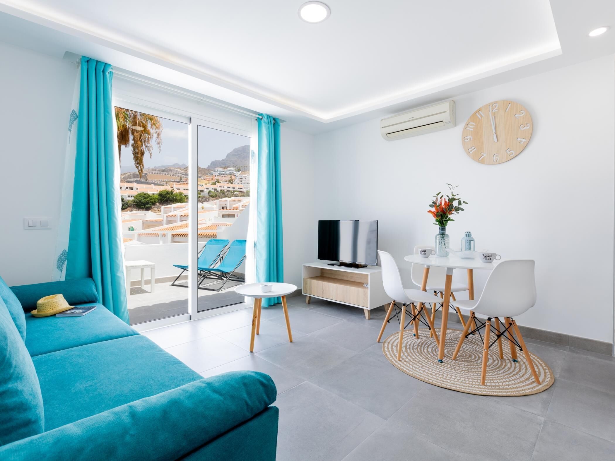 Ein modernes Wohnzimmer mit einem türkisfarbenen Sofa, einem Esstisch mit Stühlen, einem Fernseher und Blick auf eine Küstenstadt durch große Fenster.