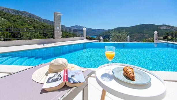 A poolside setting with a book, a croissant, and a glass of orange juice on a table, overlooking a clear blue pool and mountains.