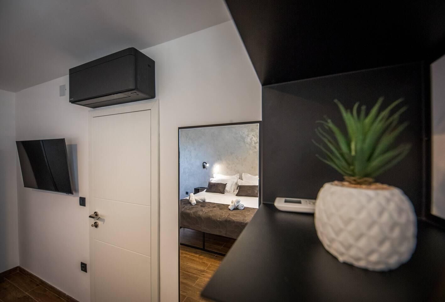 A modern hotel room with a flat-screen TV mounted on the wall, a bed with a headboard, a small bedside table with a plant, and a wall-mounted air conditioning unit.