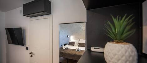 A modern hotel room with a flat-screen TV mounted on the wall, a bed with a headboard, a small bedside table with a plant, and a wall-mounted air conditioning unit.