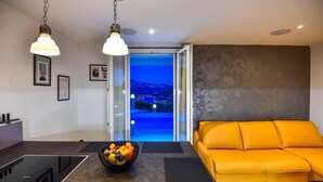 A modern living room with a yellow sofa, a dining area with a bowl of fruit, and a view of a blue-lit landscape through an open door.