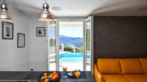 A modern living room with a large orange sofa, a dining table with fruit and drinks, and a view of a swimming pool and mountains through a glass door.
